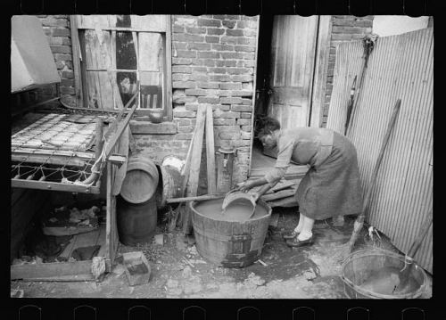 A woman collecting a bucket of water from a washbasin under a backyard water faucet, in a photo from Washington DC, originally published in 1935